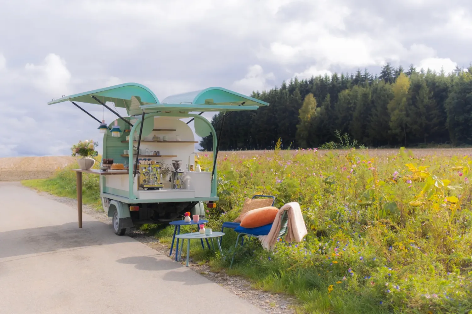 Pocissimo Coffee Ape-Mobil mit ausgeklappten Flügeln auf einem Feldweg neben blühender Wildblumenwiese